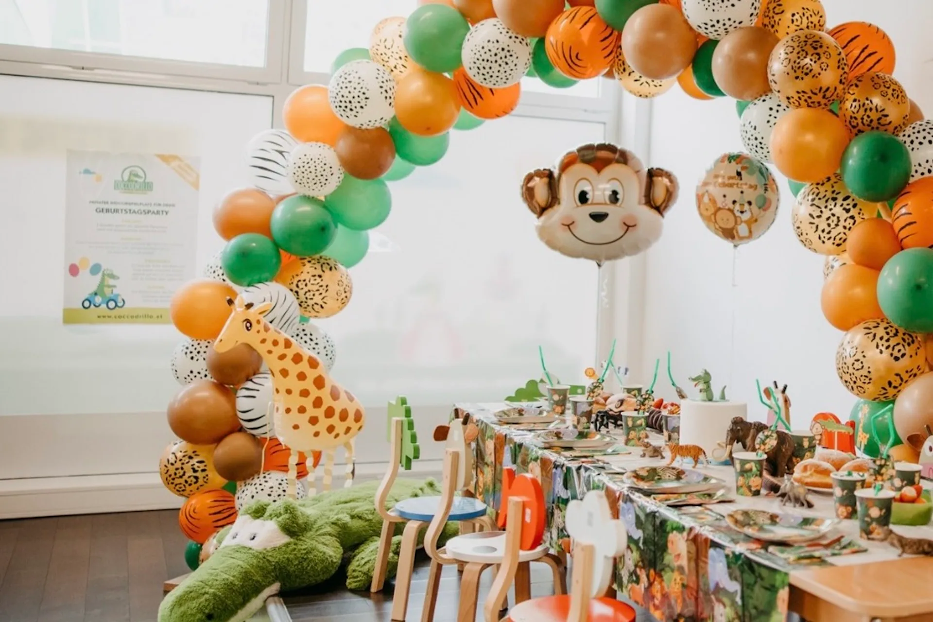 Eine Geburtstagsparty zum Thema Dschungel mit einem Bogen aus Luftballons mit Tiermotiven, einem großen Affenballon, Tierdekorationen, einer Giraffe aus Plüsch und einem Tisch mit Partygeschirr und bunten Stühlen.
