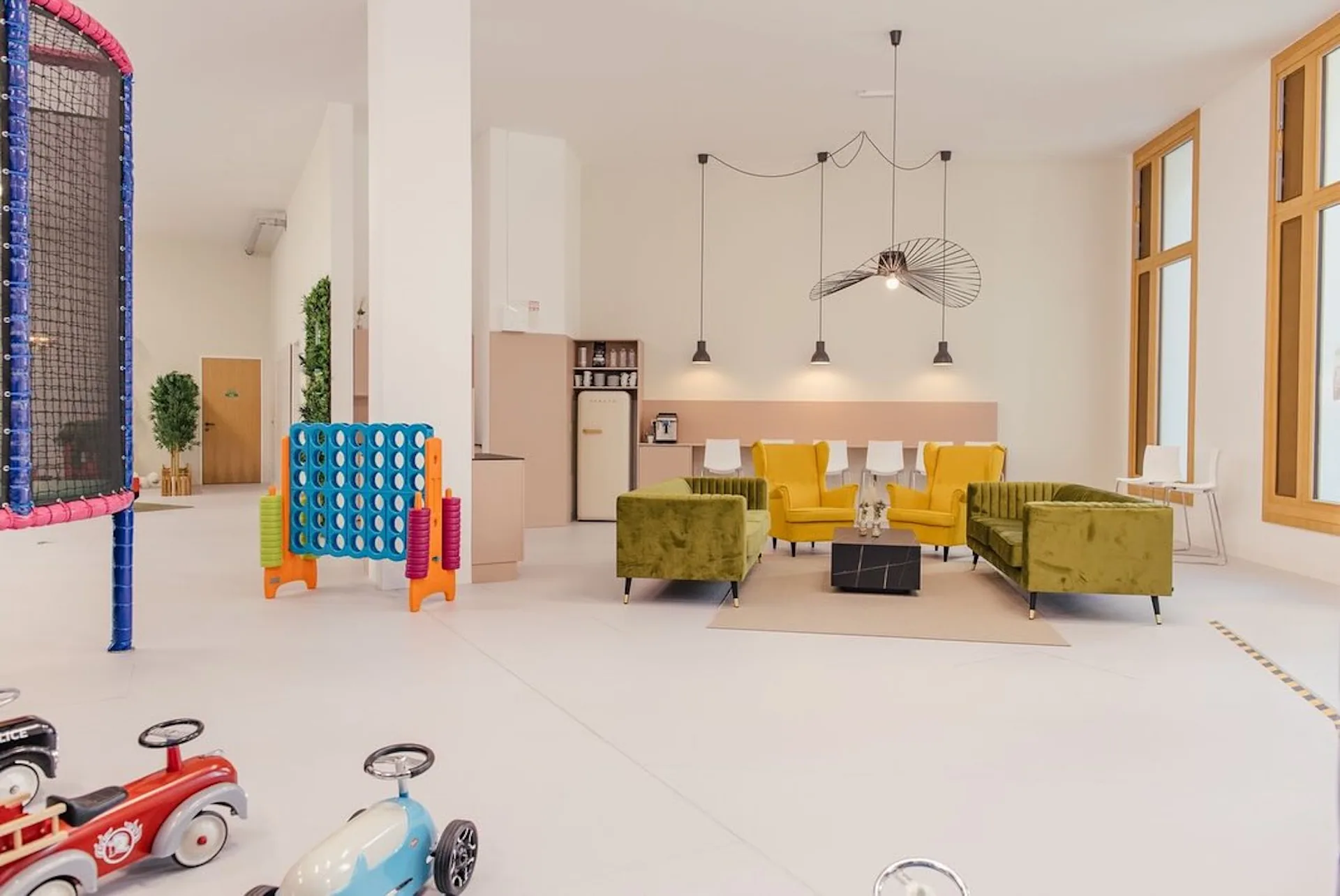 Modernes Spielzimmer mit großen Fenstern, olivgrünen und gelben Sesseln, einem Couchtisch, Hängelampen, Kinderspielzeug, einem riesigen Connect Four-Spiel und einem kleinen Klettergerüst.