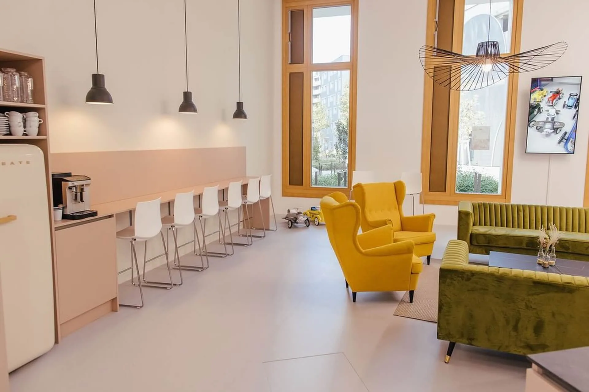 Ein modernes, helles Wohnzimmer mit hohen Fenstern, gelben Sesseln, einem grünen Samtsofa, weißen Barhockern an einem Tresen und Hängelampen. Es gibt einen Retro-Kühlschrank und eine Kaffeemaschine auf einem Seitentresen.