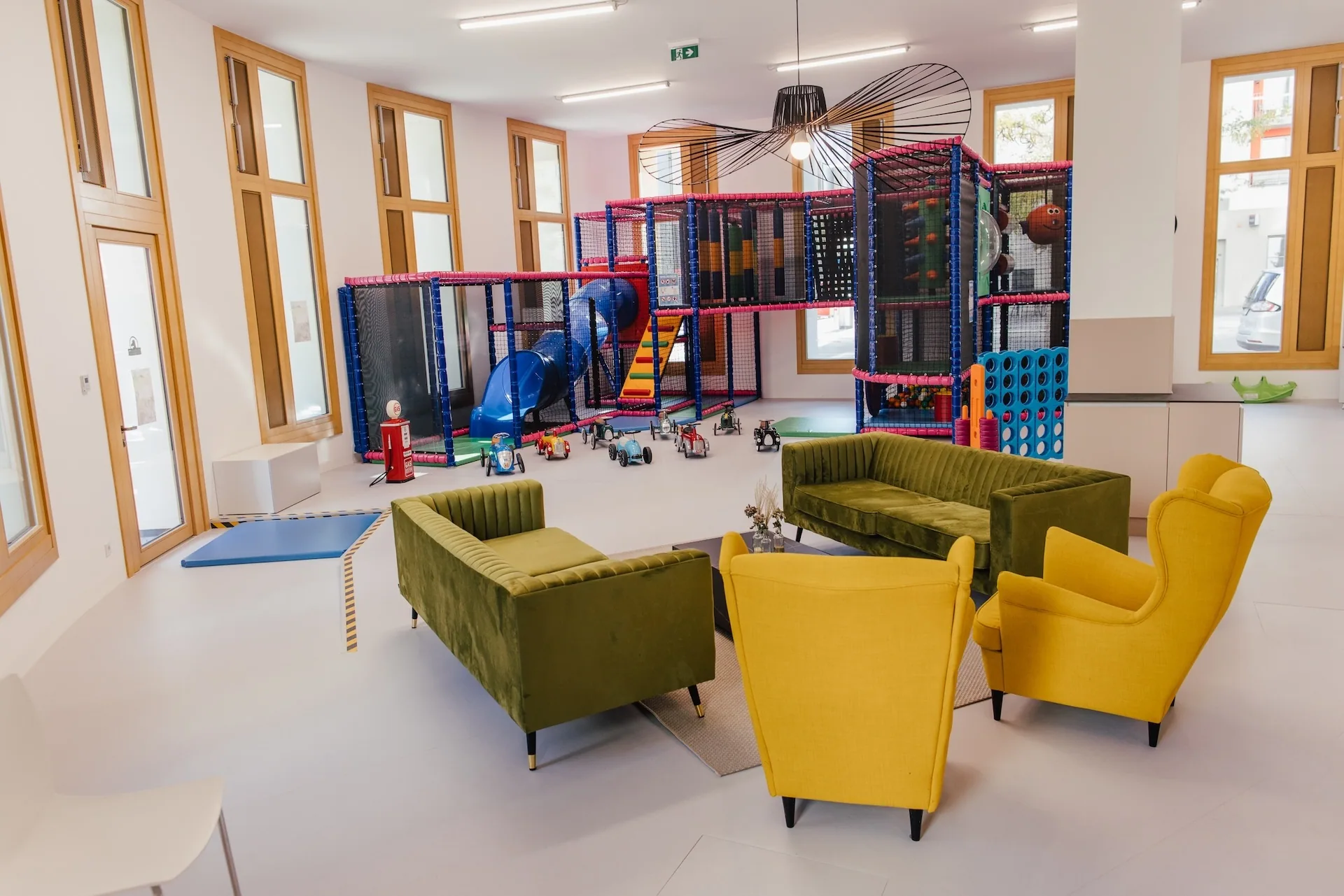 Ein modernes Spielzimmer mit großen Fenstern, grünen und gelben Sofas, Sesseln und einem Kinderspielgerät mit Rutschen, Kletternetzen und verstreuten Spielsachen auf dem Boden.