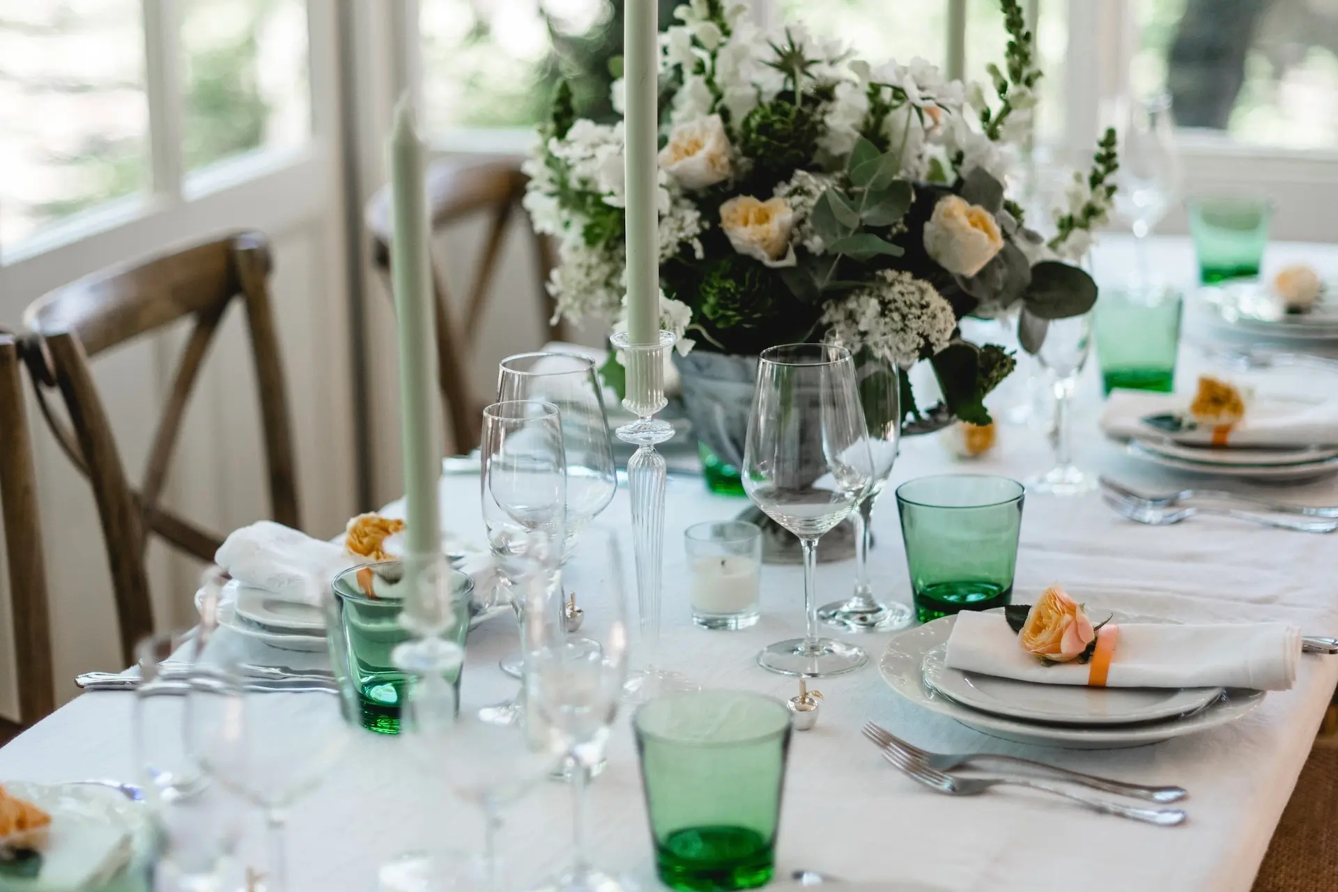 Ein schön gedeckter Esstisch mit grünen Gläsern, weißen Tellern, gefalteten Servietten, Silberbesteck, hohen Kerzen und einem Mittelstück aus weißen und pfirsichfarbenen Blumen in einem hellen Raum mit großen Fenstern.