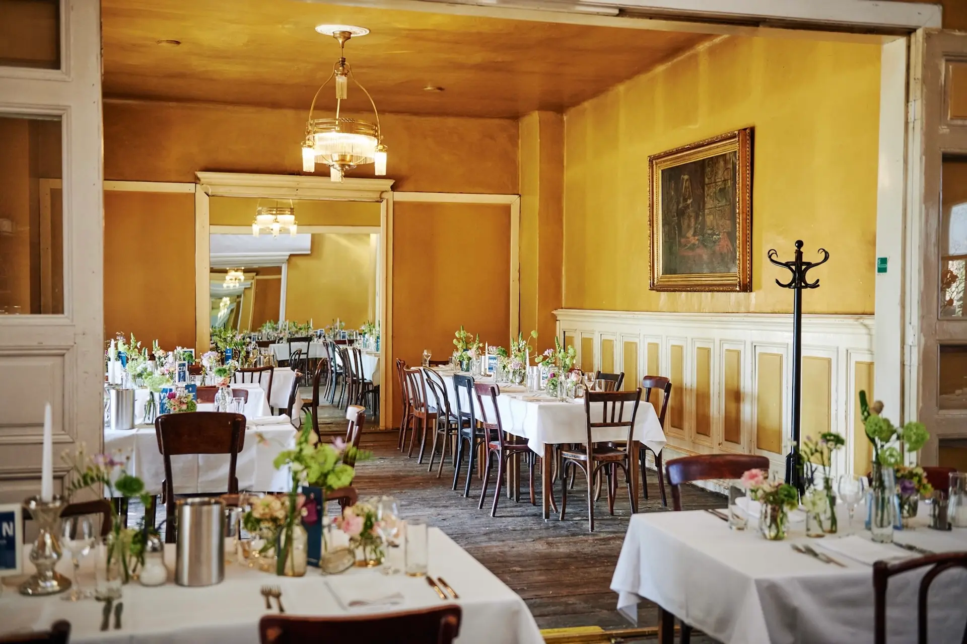 Ein warm beleuchteter Speisesaal in einem Restaurant mit gelben Wänden, Holzböden und mehreren Tischen mit weißen Tischtüchern und Blumenkränzen. Stühle umgeben die Tische, und an der Wand hängt ein gerahmtes Gemälde.