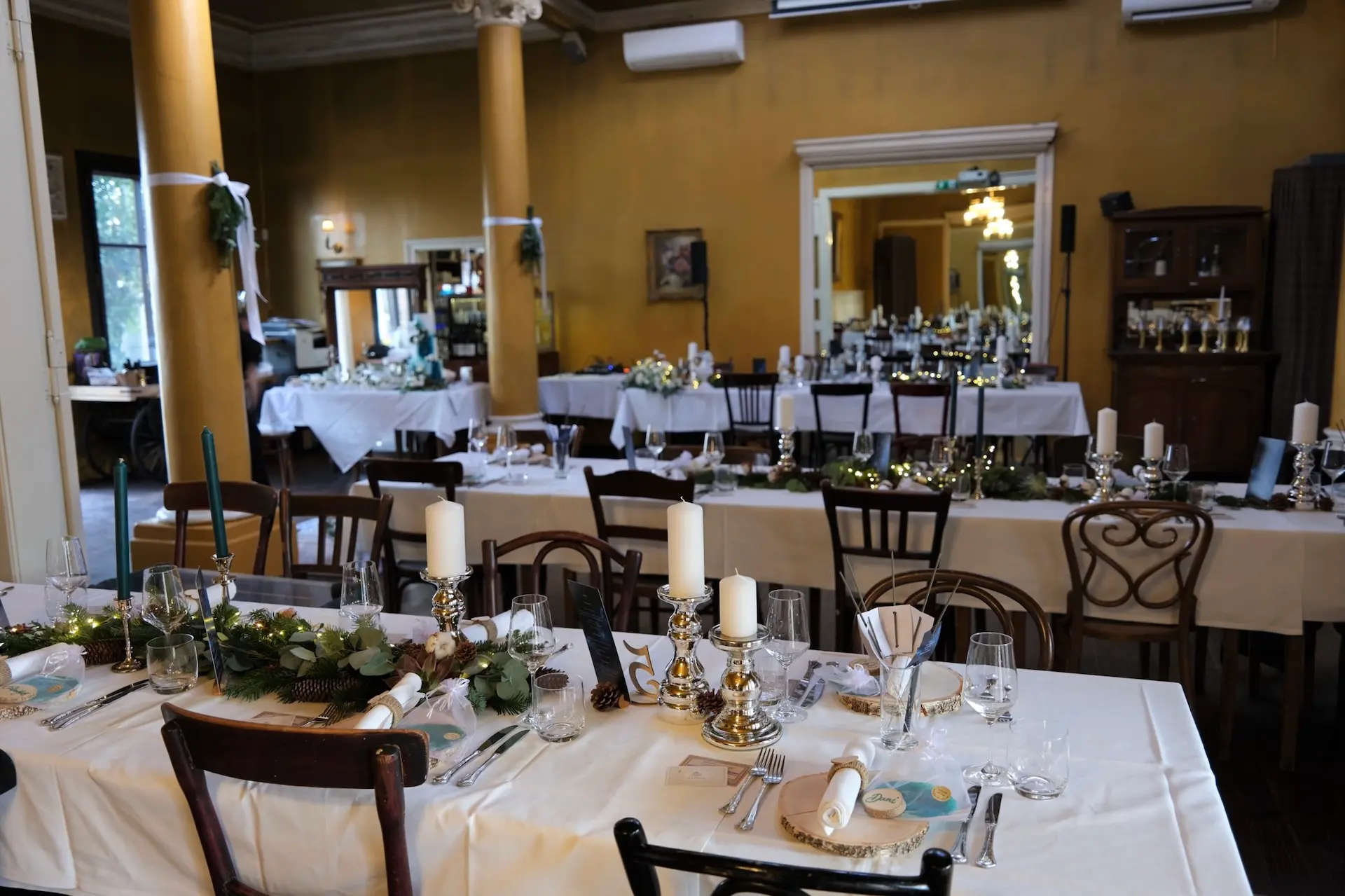 Elegantes Esszimmer für eine Veranstaltung mit weißen Tischtüchern, Kerzen, grünen Tafelaufsätzen und Gedecken auf langen Tischen; goldene Säulen und große Spiegel im Hintergrund spiegeln den dekorierten Raum wider.