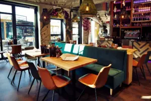 Ein modernes Restaurantinterieur mit Holztischen, grün gepolsterten Sitznischen und hellbraunen Stühlen. Sonnenlicht strömt durch große Fenster, während bunte Wandkunst und stilvolle Pendelleuchten dieses Beispiel für coole Restaurants Wien aufwerten.