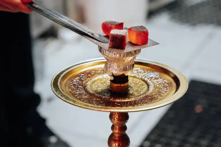 Nahaufnahme einer Wasserpfeife mit glühenden Holzwürfeln, die auf einen Metallschirm über der mit Folie bedeckten Schale gelegt werden. Der verschnörkelte goldene Sockel der Wasserpfeife ist sichtbar.