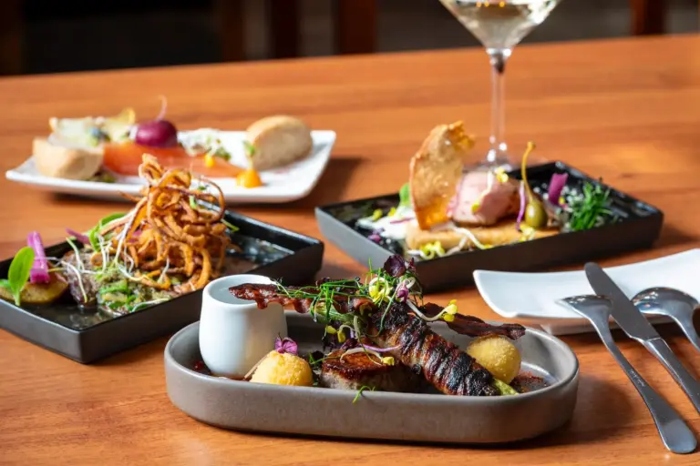 Drei Feinschmeckergerichte sind kunstvoll auf einem Holztisch angerichtet, jedes mit bunten Garnituren und kunstvoller Präsentation. Im Hintergrund steht ein Glas Weißwein neben sauber arrangiertem Silberbesteck und einem weißen, rechteckigen Teller.