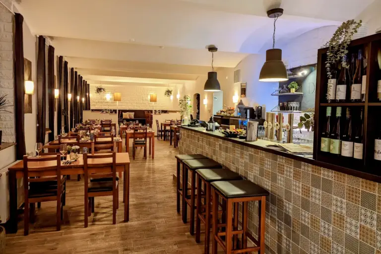 Ein gemütliches Restaurant mit Holztischen und -stühlen, einem gefliesten Bartresen, Hängelampen, Weinflaschen in Regalen und warmer Umgebungsbeleuchtung. Der Raum wirkt einladend und ordentlich für die Gäste hergerichtet.