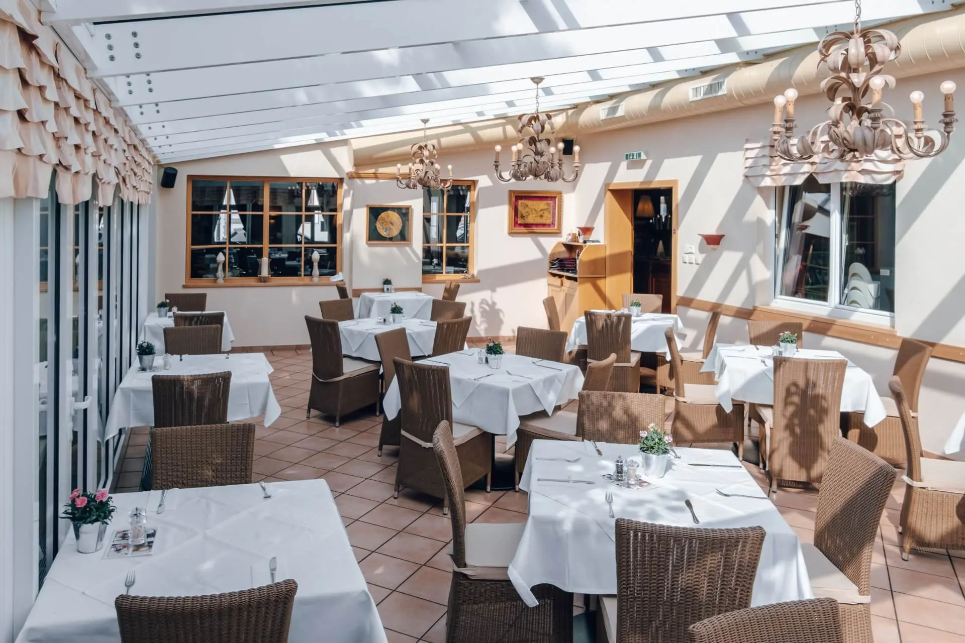 Ein heller, sonnenbeschienener Restaurantbereich mit Korbstühlen und Tischen mit weißen Tischdecken. Von der Decke hängen Kronleuchter, und große Fenster lassen viel natürliches Licht herein. Kleine Blumenarrangements schmücken jeden Tisch.