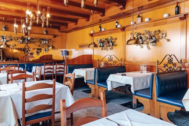 Ein gemütliches Restaurant mit hölzernen Wänden und Decken, blauen gepolsterten Bänken, Holzstühlen, weißen Tischdecken und Weinflaschen, die in dekorativen Regalen an den Wänden stehen. Von der Decke hängen Kronleuchter.