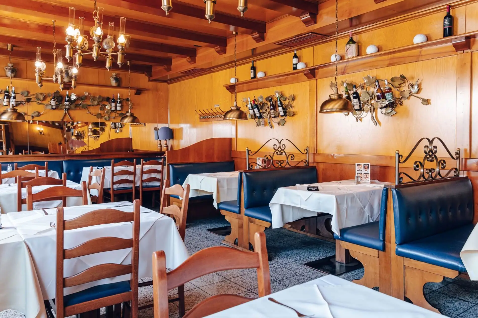 Ein gemütliches Restaurant mit hölzernen Wänden und Decken, blauen gepolsterten Bänken, Holzstühlen, weißen Tischdecken und Weinflaschen, die in dekorativen Regalen an den Wänden stehen. Von der Decke hängen Kronleuchter.