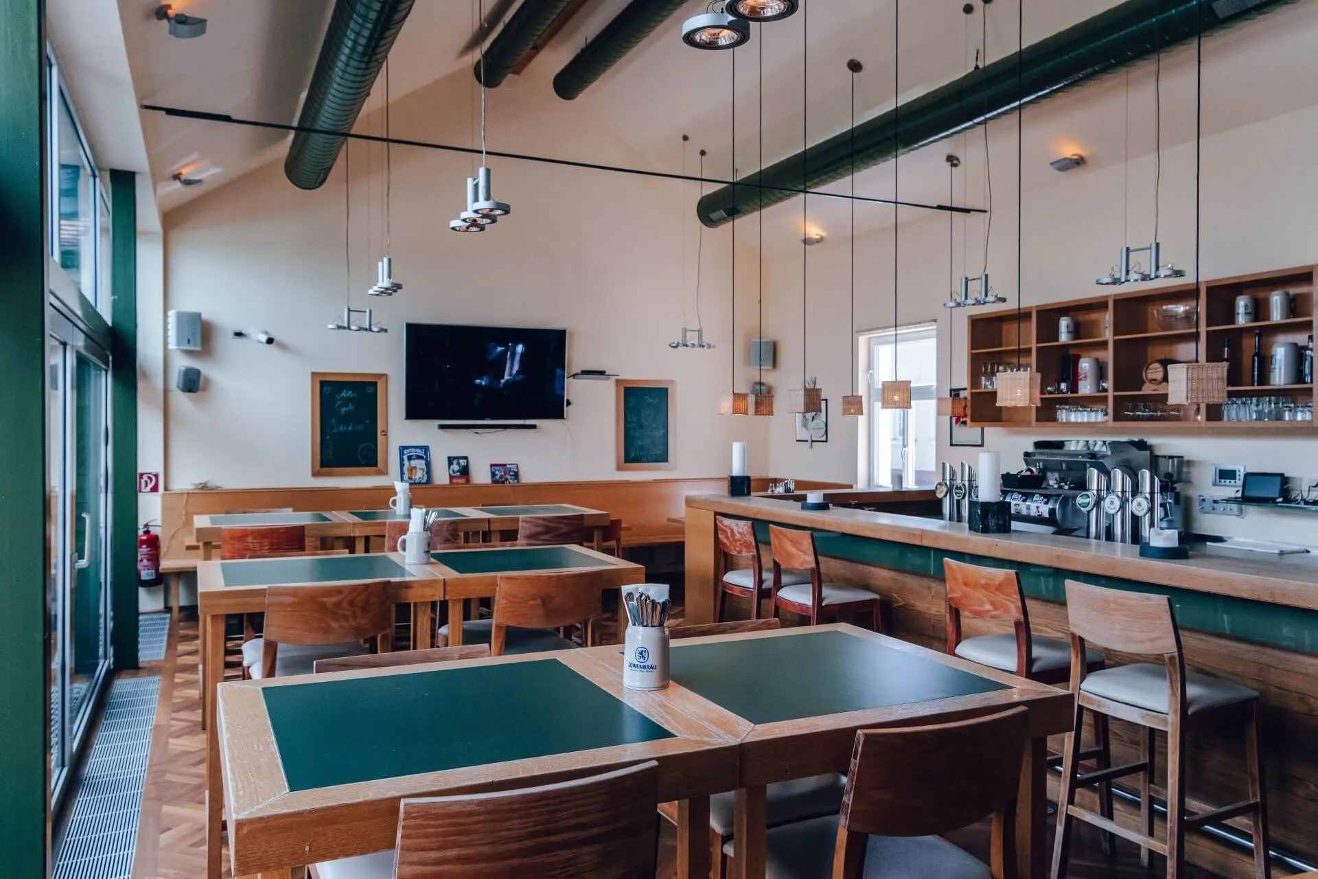 Modernes Restaurantinterieur mit Holztischen und -stühlen, grünen Tischplatten, einer langen Bar mit Hockern, Hängelampen, einem an der Wand montierten Fernseher, Regalen mit Gläsern und großen Fenstern, die natürliches Licht hereinlassen.