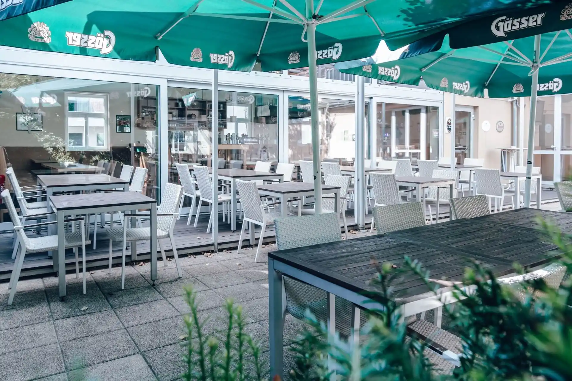 Außensitzbereich eines Cafés oder Restaurants mit leeren weißen Tischen und Stühlen unter großen grünen Sonnenschirmen, neben einem Gebäude mit Glaswänden. Im Vordergrund sind einige Pflanzen zu sehen.