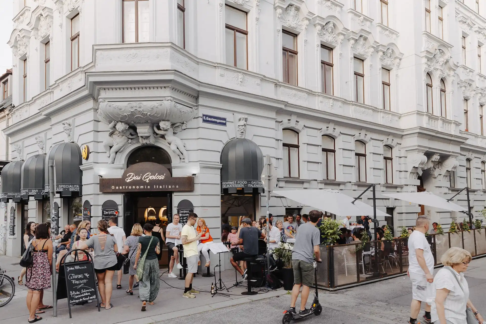 Eine belebte Straßenszene vor einem weißen, verschnörkelten Gebäude mit Menschen, die sich in einem Café und Restaurant versammelt haben, einige sitzen unter Sonnenschirmen, andere stehen oder gehen auf dem Bürgersteig vorbei.
