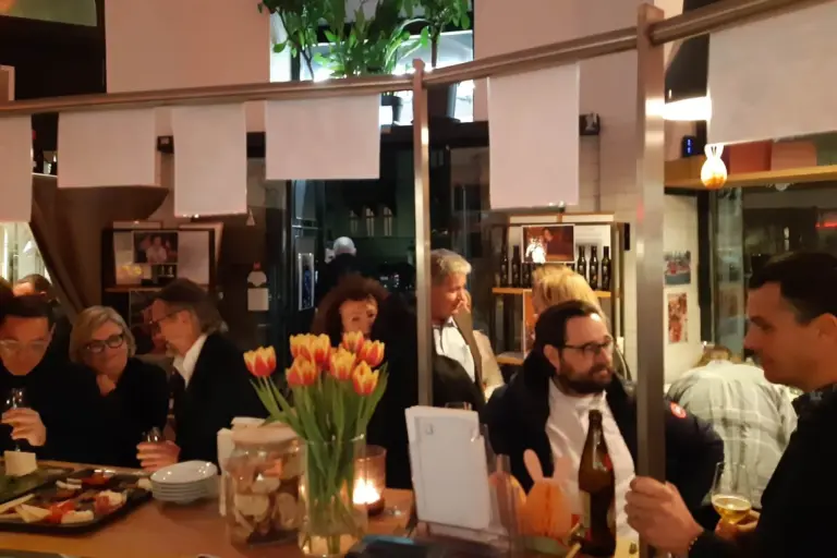 Eine Gruppe von Menschen sitzt und steht um eine Bar in einem gemütlichen Restaurant, plaudert und genießt Getränke. Auf der Bar stehen Tulpen, Kerzen und Snacks, und im Hintergrund stehen Weinflaschen in Regalen. Die Atmosphäre wirkt lebendig und warm.