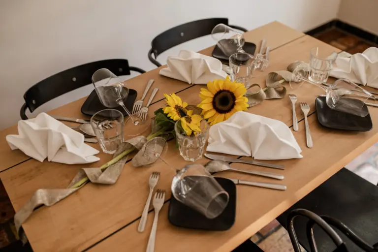 Ein hölzerner Esstisch für vier Personen mit schwarzen Tellern, Gläsern, Silberbesteck, weißen Servietten, die dekorativ gefaltet sind, und einem Mittelstück aus gelben Sonnenblumen und einem goldenen Band. Vier schwarze Stühle umgeben den Tisch.
