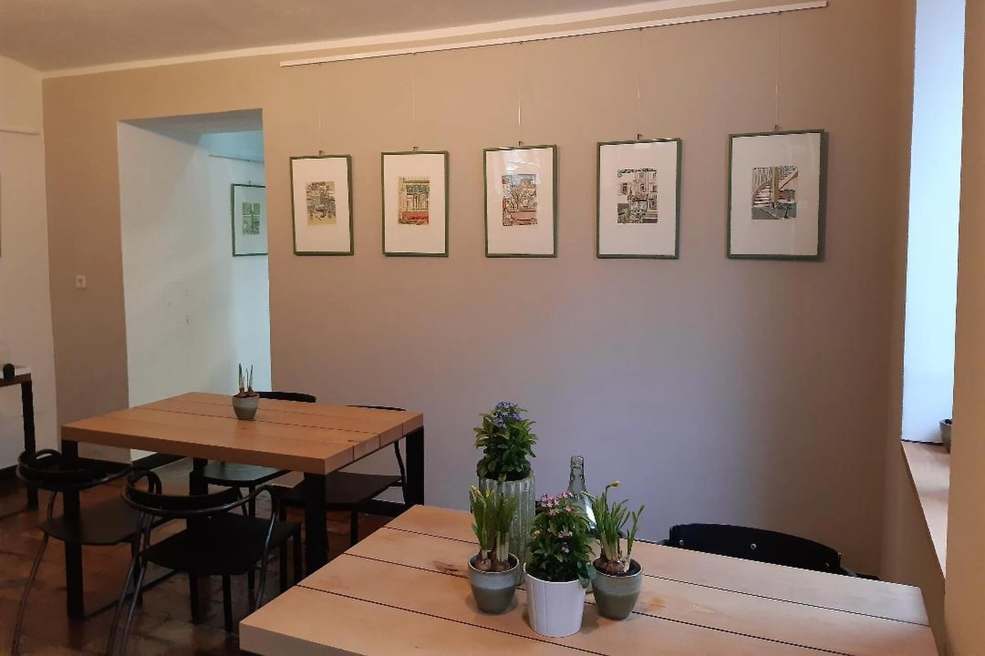 Ein kleiner, gemütlicher Raum mit hellen Holztischen, schwarzen Stühlen und Topfpflanzen auf den Tischen. Sechs gerahmte Kunstwerke hängen in einer Reihe an der beigen Wand. Von der rechten Seite fällt natürliches Licht ein.
