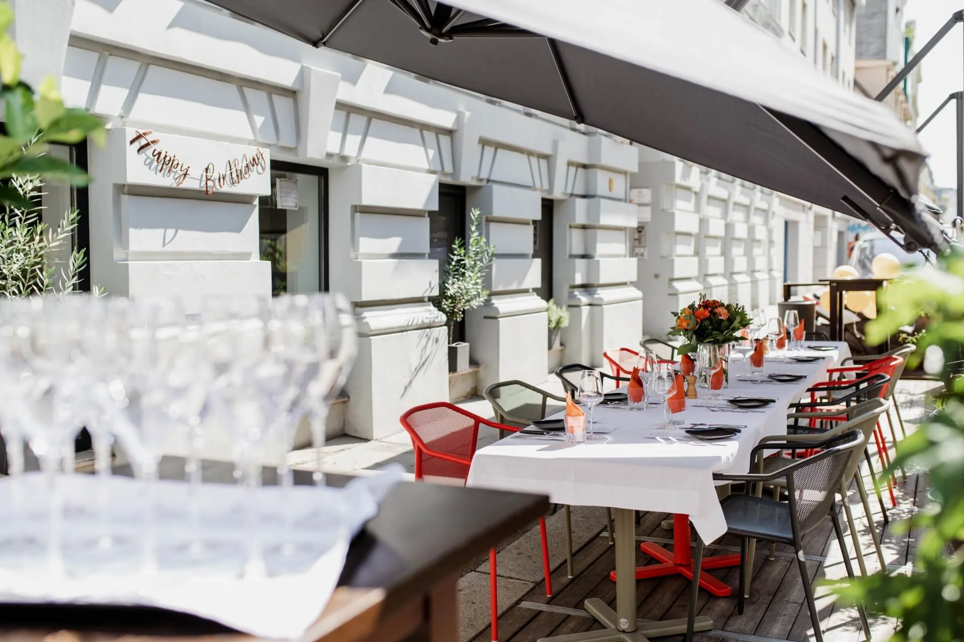 Ein langer, für eine Feier gedeckter Tisch im Freien mit weißen Tischtüchern, orangefarbenen Getränken, Blumen und Gedecken; ein "Happy Birthday"-Banner hängt an einer Gebäudewand, mit Stühlen und Gläsern unter einem großen schwarzen Sonnenschirm.