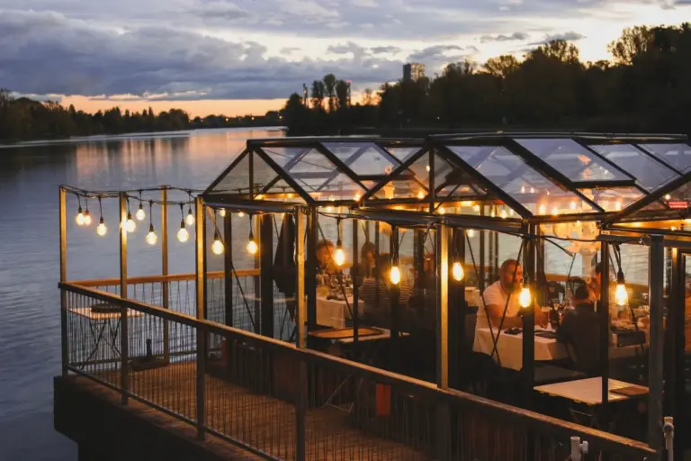 Ein gläsernes Restaurant am Flussufer erstrahlt in der Abenddämmerung mit Lichterketten; die Gäste sitzen an Tischen im Inneren, während im Hintergrund das ruhige Wasser und die Bäume unter dem bewölkten Abendhimmel zu sehen sind.