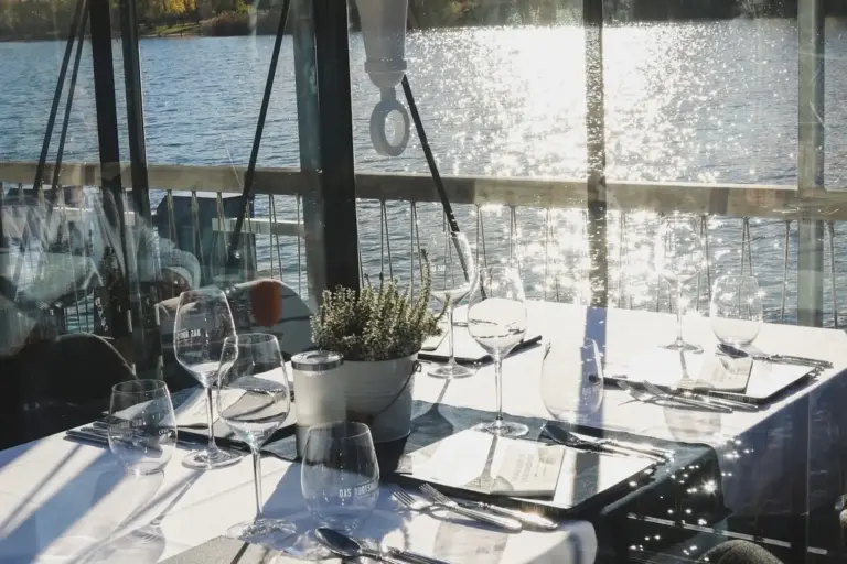 Ein sonnendurchfluteter Restauranttisch mit Gläsern, Tellern und einer Topfpflanze blickt durch große Fenster auf einen glitzernden See und schafft eine ruhige und elegante Essatmosphäre.