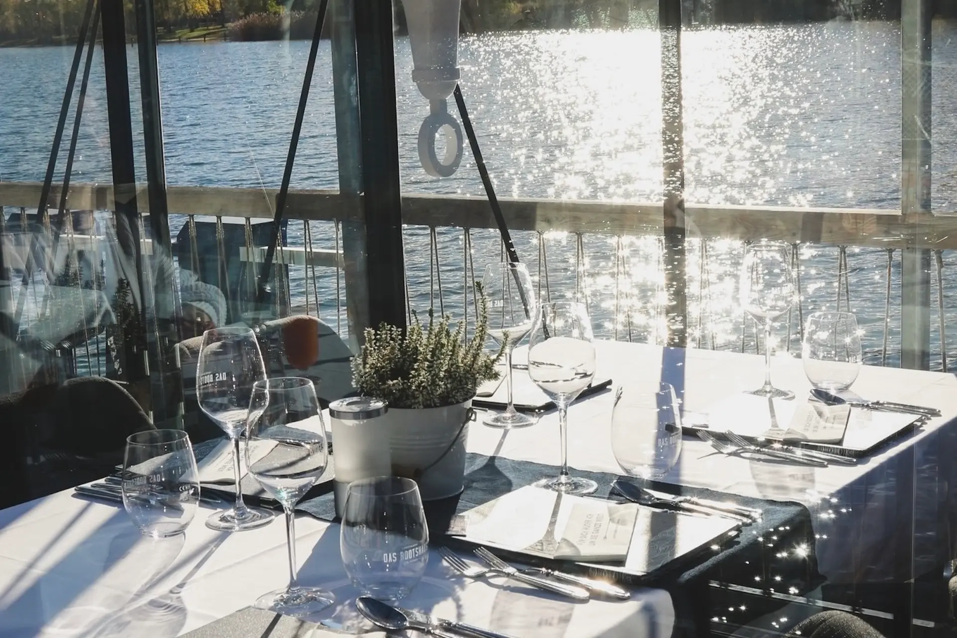 Ein sonnendurchfluteter Restauranttisch mit Gläsern, Tellern und einer Topfpflanze blickt durch große Fenster auf einen glitzernden See und schafft eine ruhige und elegante Essatmosphäre.