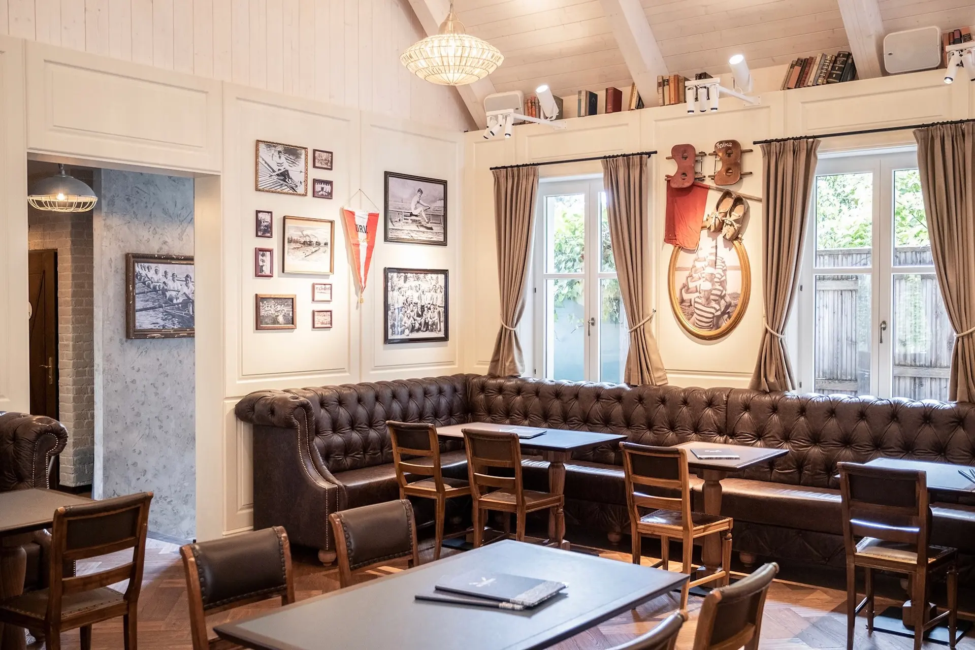 Ein gemütliches, helles Café mit braunen Ledersesseln, Holzstühlen und -tischen, gerahmten Fotos und Erinnerungsstücken an cremefarbenen Wänden, zwei großen Fenstern mit Vorhängen und Büchern in einem Regal darüber.