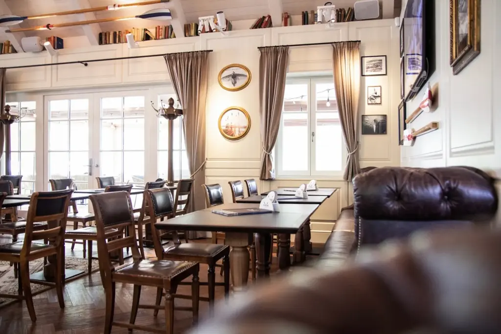 Ein gemütliches, sonnendurchflutetes Restaurant mit Holztischen und -stühlen, Ledersesseln, großen Fenstern mit Vorhängen und Dekoration zum Thema Luftfahrt an den Wänden, darunter Modellflugzeuge und gerahmte Bilder.