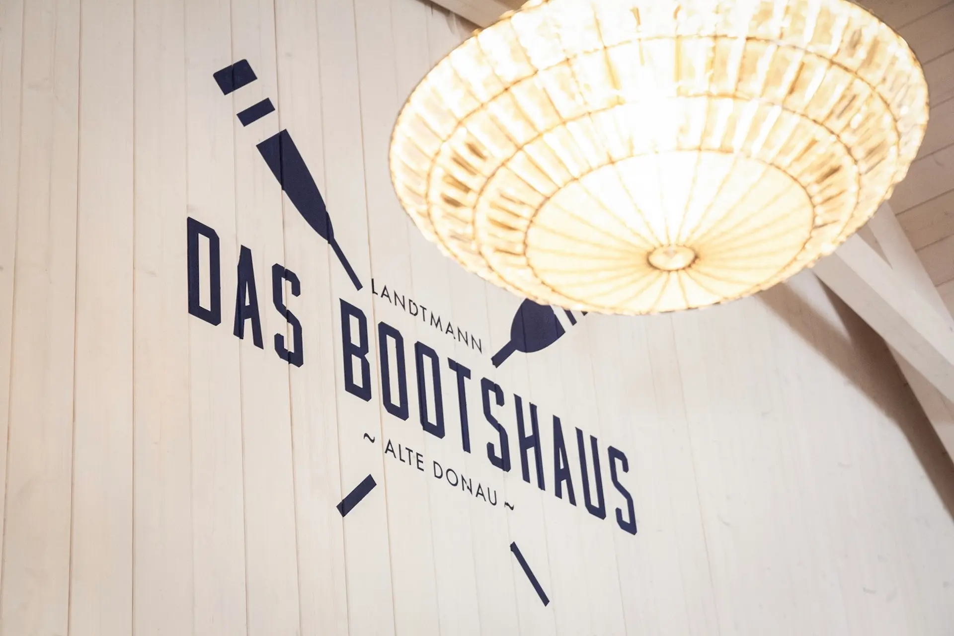 Eine weiße Wand mit gekreuzten Rudern und der Aufschrift "Das Bootshaus Landtmann Alte Donau" in fetten, marineblauen Buchstaben, darunter eine große, leuchtende, runde, geflochtene Leuchte.