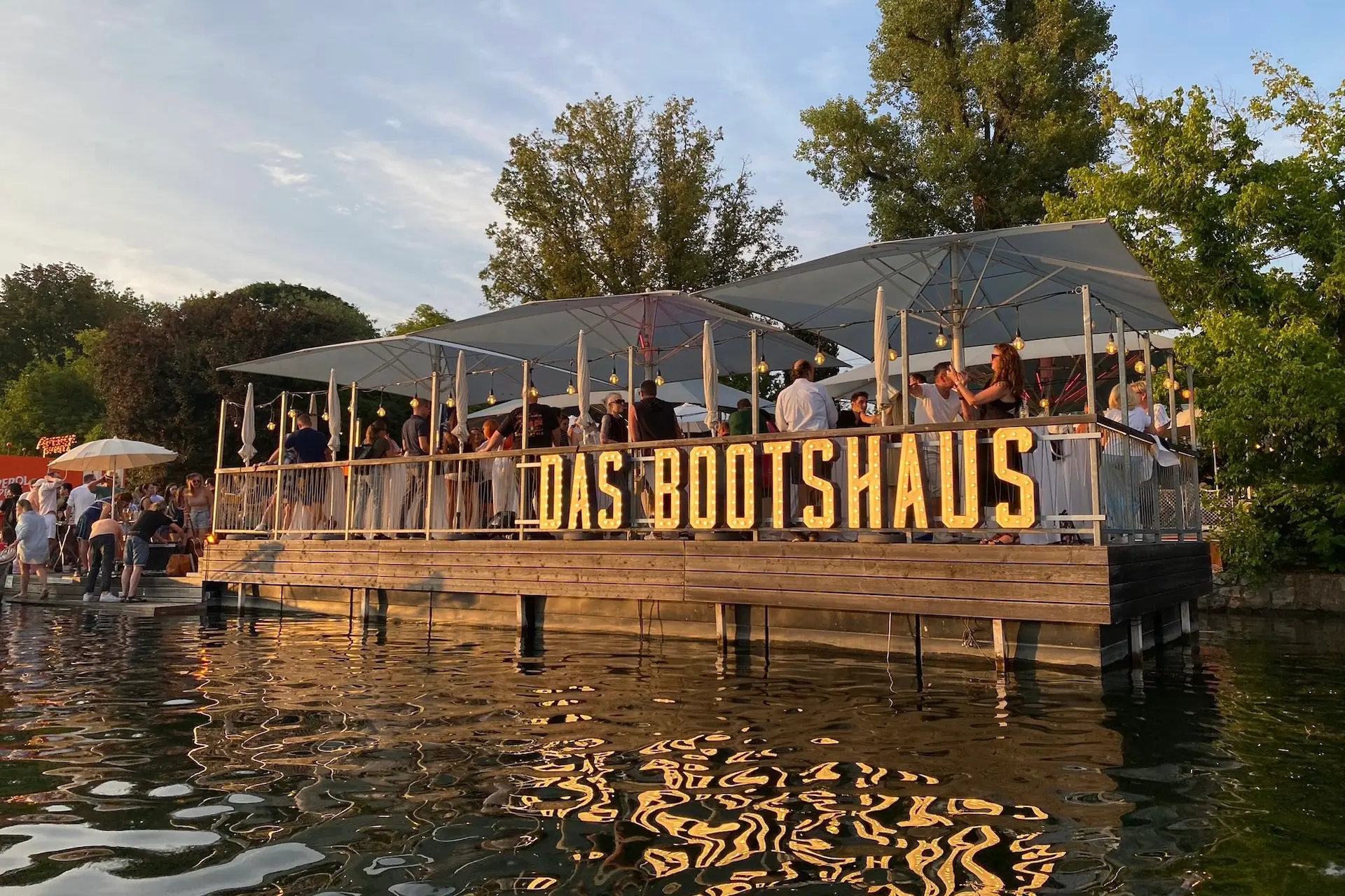 Eine schwimmende Holzbar namens Das Bootshaus ist überfüllt mit Menschen unter einem Baldachin, umgeben von Bäumen, und spiegelt sich bei Sonnenuntergang im ruhigen Wasser.