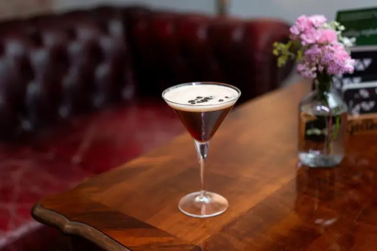 Ein Martiniglas, gefüllt mit einem cremefarbenen Cocktail, garniert mit Kaffeebohnen, steht auf einem polierten Holztisch neben einer kleinen Vase mit rosa Blumen. Im Hintergrund steht ein rotes Ledersofa.