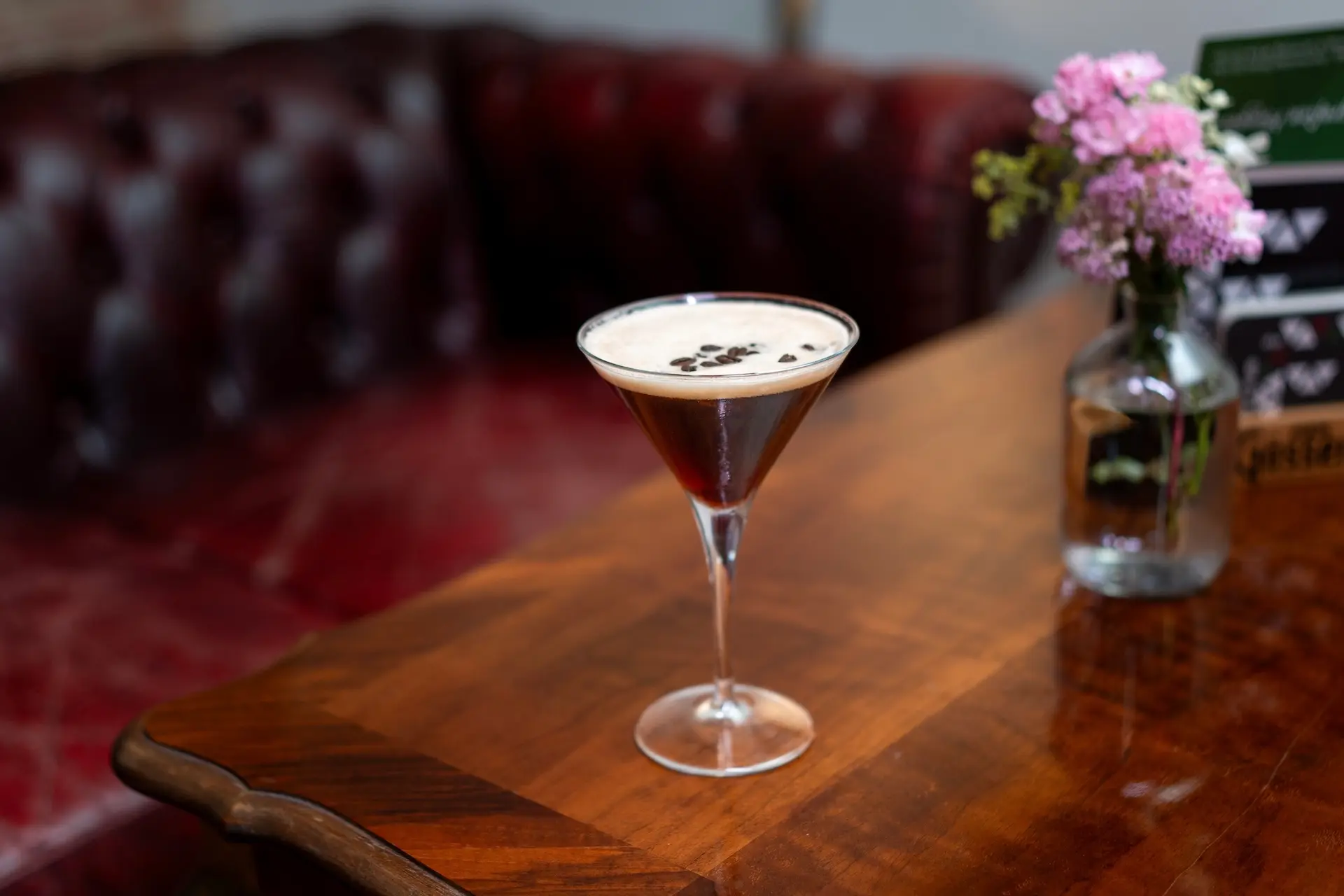 Ein Martiniglas, gefüllt mit einem cremefarbenen Cocktail, garniert mit Kaffeebohnen, steht auf einem polierten Holztisch neben einer kleinen Vase mit rosa Blumen. Im Hintergrund steht ein rotes Ledersofa.