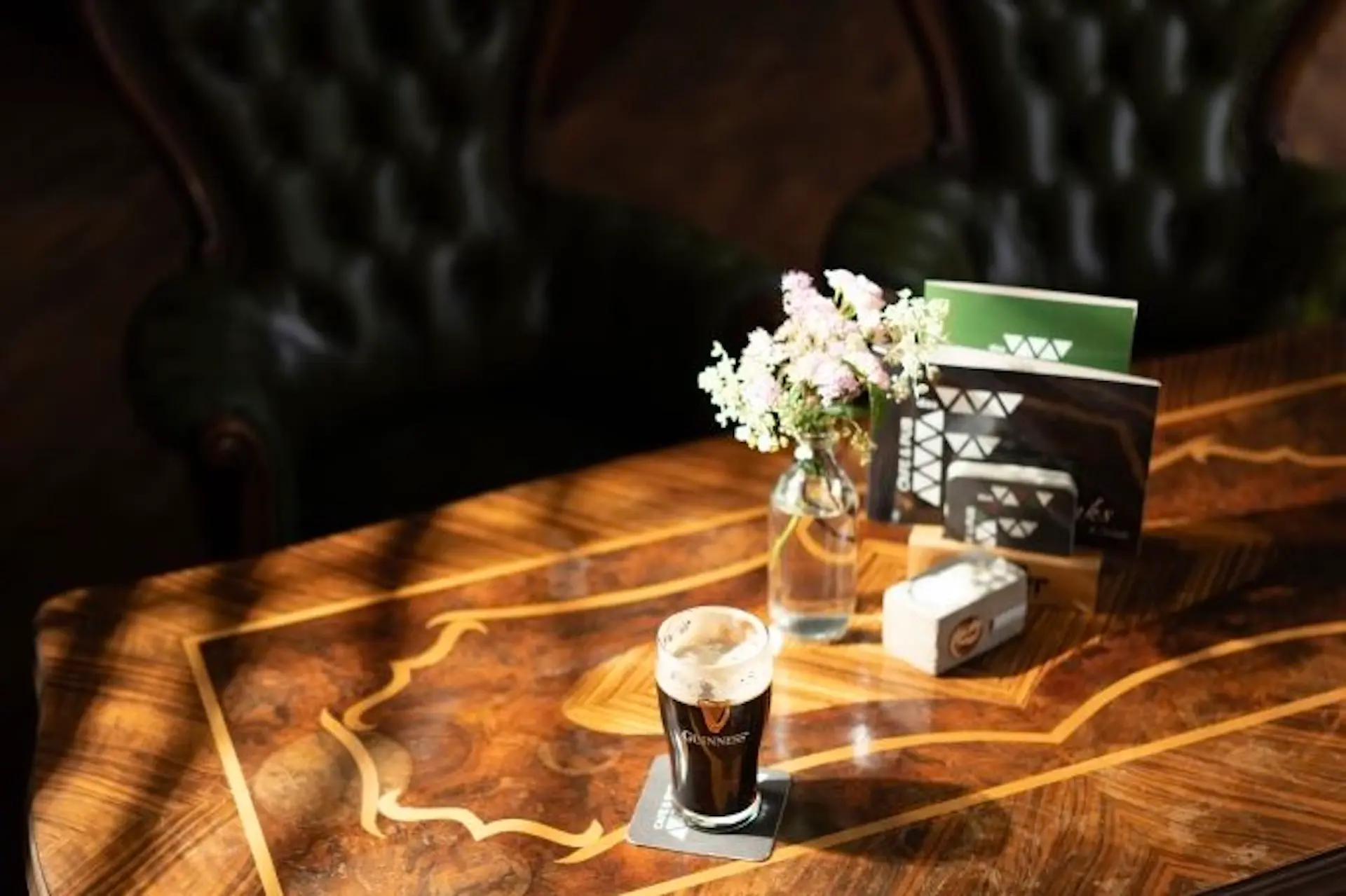 Ein dunkles Bier mit cremiger Schaumkrone steht auf einem Untersetzer auf einem polierten Holztisch. Daneben stehen eine kleine Blumenvase, Speisekarten und ein Serviettenhalter. Im Hintergrund stehen grüne, getuftete Stühle.
