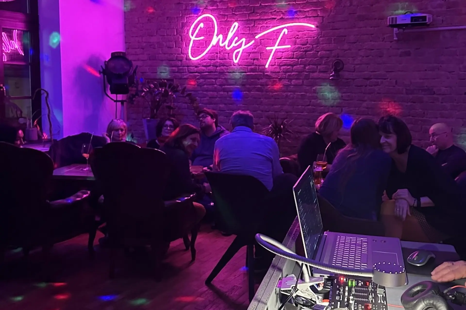 Das F Menschen sitzen und unterhalten sich in einer schwach beleuchteten Bar mit violetter Beleuchtung. Auf einem Neonschild an einer freiliegenden Backsteinmauer steht "Only F". Im Vordergrund ist eine Tonanlage zu sehen, und bunte Lichtpunkte zieren die Szene.
