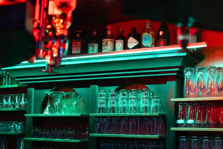 Die Regale der Bar sind mit rotem und grünem Neonlicht beleuchtet. Auf dem obersten Regal stehen verschiedene Spirituosenflaschen, während auf den unteren Regalen mehrere Reihen von Gläsern, darunter Weingläser und Becher, übersichtlich angeordnet sind.