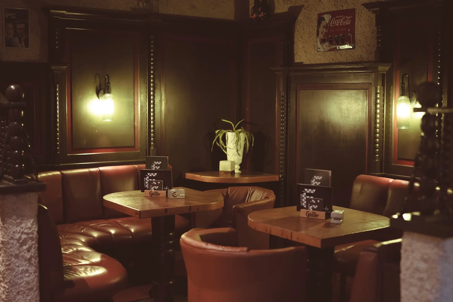 Ein gemütliches, schwach beleuchtetes Café oder Pub mit roten Ledersitzen, Holztischen und warmen Wandlampen. Ein weißer Blumentopf steht auf einem Sims in der Mitte, und alte Schilder und Speisekarten schmücken die dunkel getäfelten Wände.