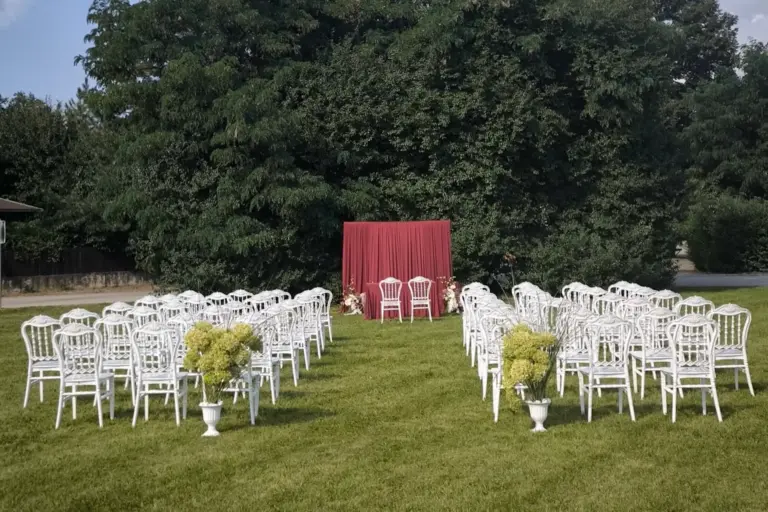 Hochzeit im Freien mit Reihen von weißen Stühlen vor einem roten Hintergrund und zwei Stühlen an der Vorderseite, alles auf grünem Gras mit Bäumen im Hintergrund und Topfpflanzen am Gang.