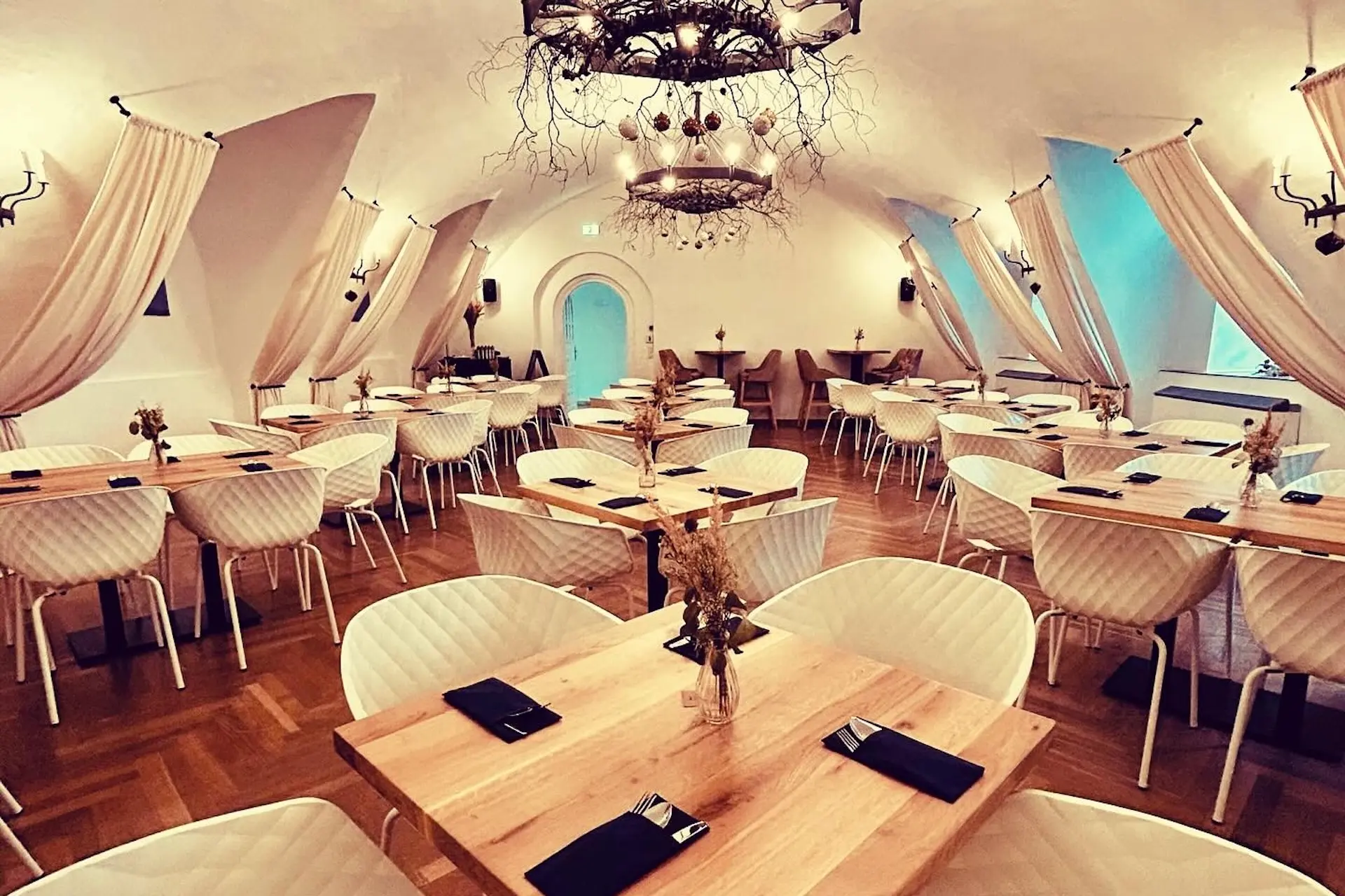 Ein stilvolles Restaurant mit gewölbten Decken, modernen Kronleuchtern, hellen Holztischen, weißen gepolsterten Stühlen und eleganten Vorhängen. Die Tische sind mit schwarzen Servietten und getrockneten Blumensträußen gedeckt.