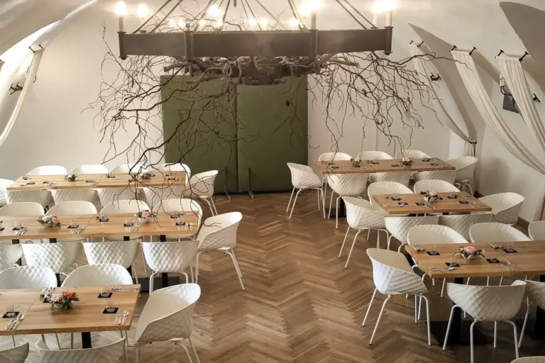 Ein moderner Speisesaal mit Holztischen und weißen Stühlen, die für eine Mahlzeit gedeckt sind. Ein großer Kronleuchter mit kahlen Ästen hängt von der Decke über den Tischen. Der Boden hat ein Fischgrätenmuster.