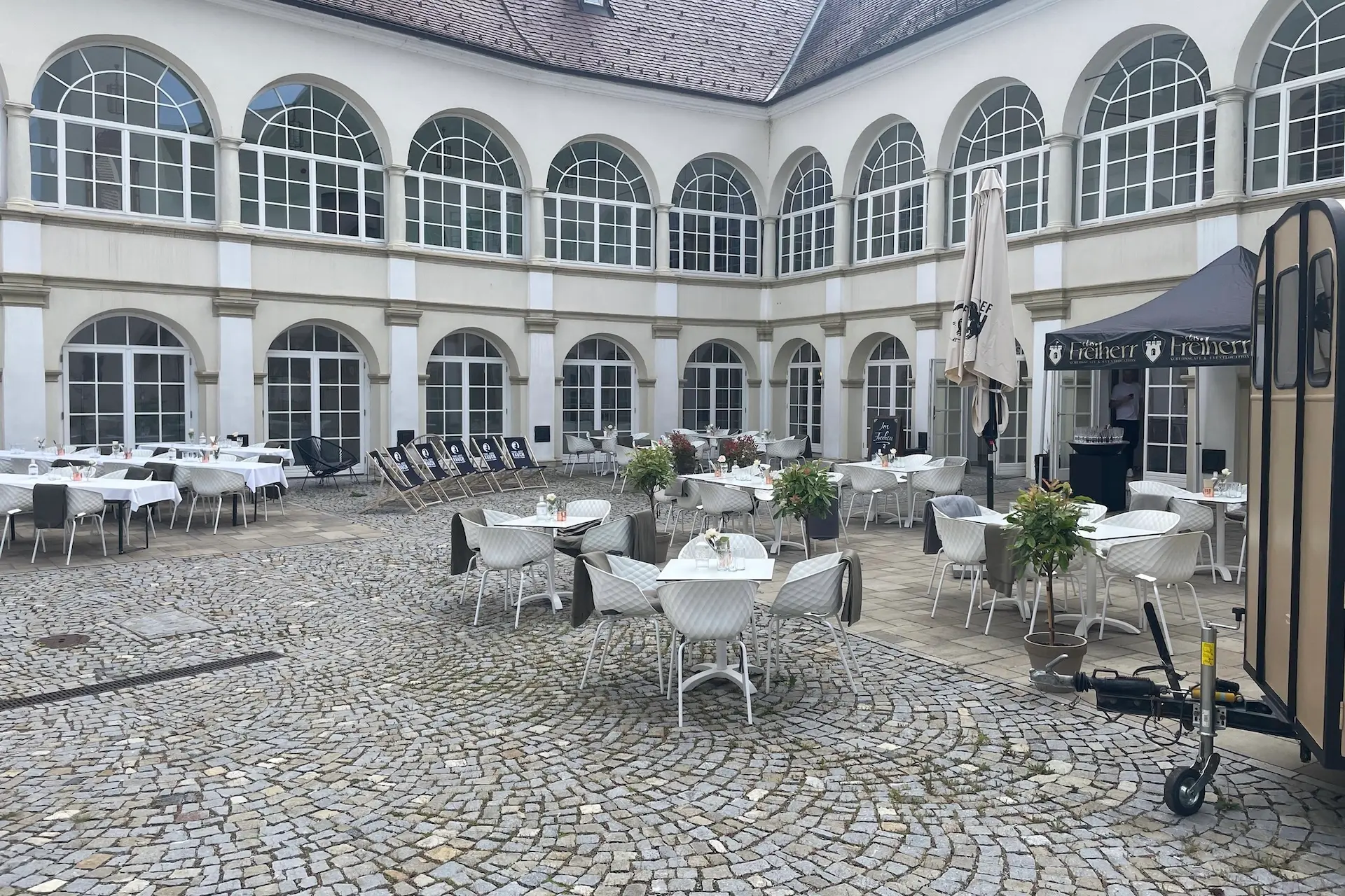 Das Freiherr In einem Innenhof mit Kopfsteinpflaster stehen weiße Tische und Stühle zum Essen bereit. Große gewölbte Fenster säumen das umliegende Gebäude, und Topfpflanzen schmücken den Raum. Einige leere Liegestühle sind an der Wand aufgestellt.