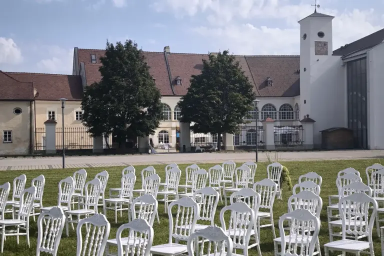 Vor einem historischen Gebäude mit Bogenfenstern, einem Uhrenturm und Bäumen stehen bei teilweise bewölktem Himmel Reihen leerer weißer Stühle im Gras.