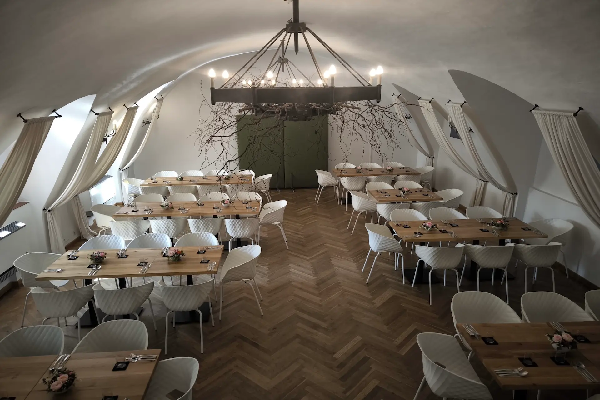 Ein geräumiger Speisesaal mit Holztischen, weißen Stühlen und Blumenkränzen. Ein großer Kronleuchter mit Zweigen hängt von der gewölbten Decke, und Vorhänge umrahmen die Wände. Der Raum ist ordentlich eingerichtet und gut beleuchtet.