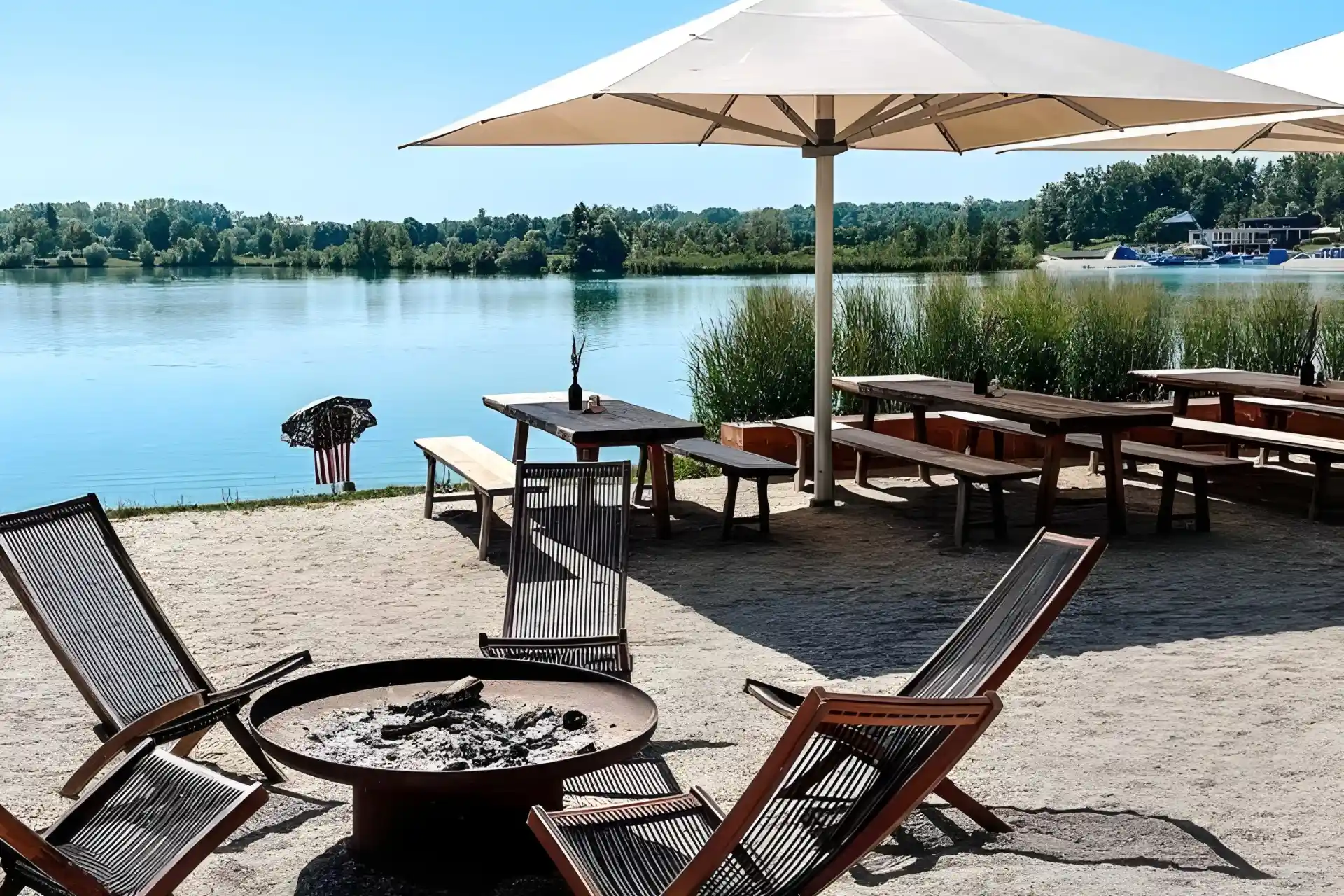 Sitzbereich im Freien am See mit hölzernen Liegestühlen um eine Feuerstelle, Picknicktischen unter einem großen weißen Sonnenschirm und Grünpflanzen im Hintergrund an einem sonnigen Tag.
