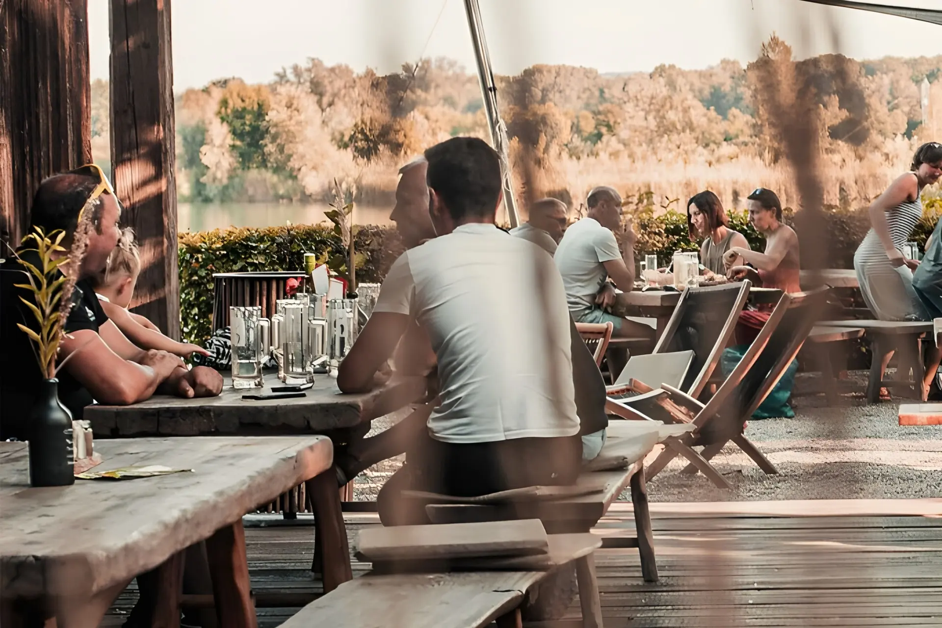 Die Menschen sitzen an Holztischen auf einer Terrasse im Freien, essen und unterhalten sich in einer entspannten, sonnenbeschienenen Umgebung mit Bäumen und Grünpflanzen im Hintergrund. Gläser und Teller stehen auf den Tischen, und einige Leute stehen sich gegenüber und unterhalten sich.
