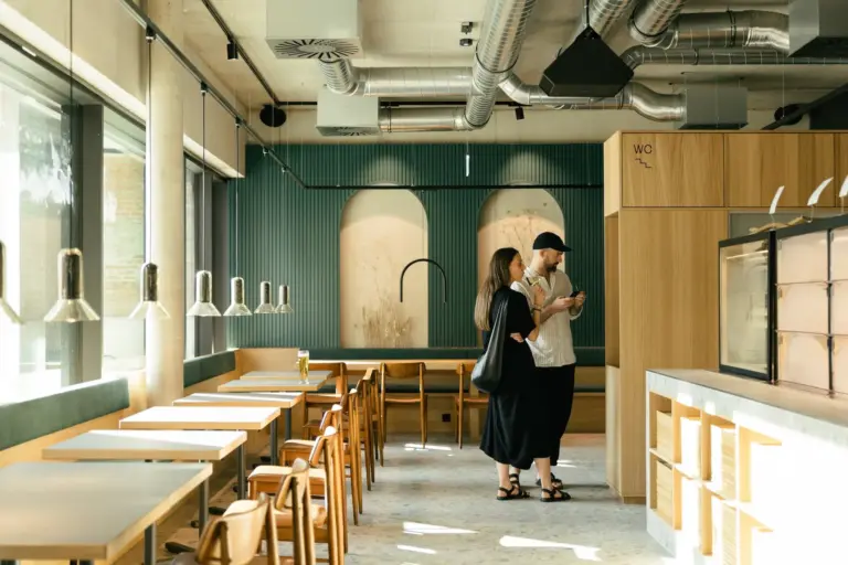 Ein Paar steht in einem modernen, größtenteils leeren Café mit Holztischen und -stühlen, grünen Wänden, großen Fenstern und industriellen Deckenlüftern zusammen. Der Mann schaut auf sein Telefon, während die Frau dicht neben ihm steht.