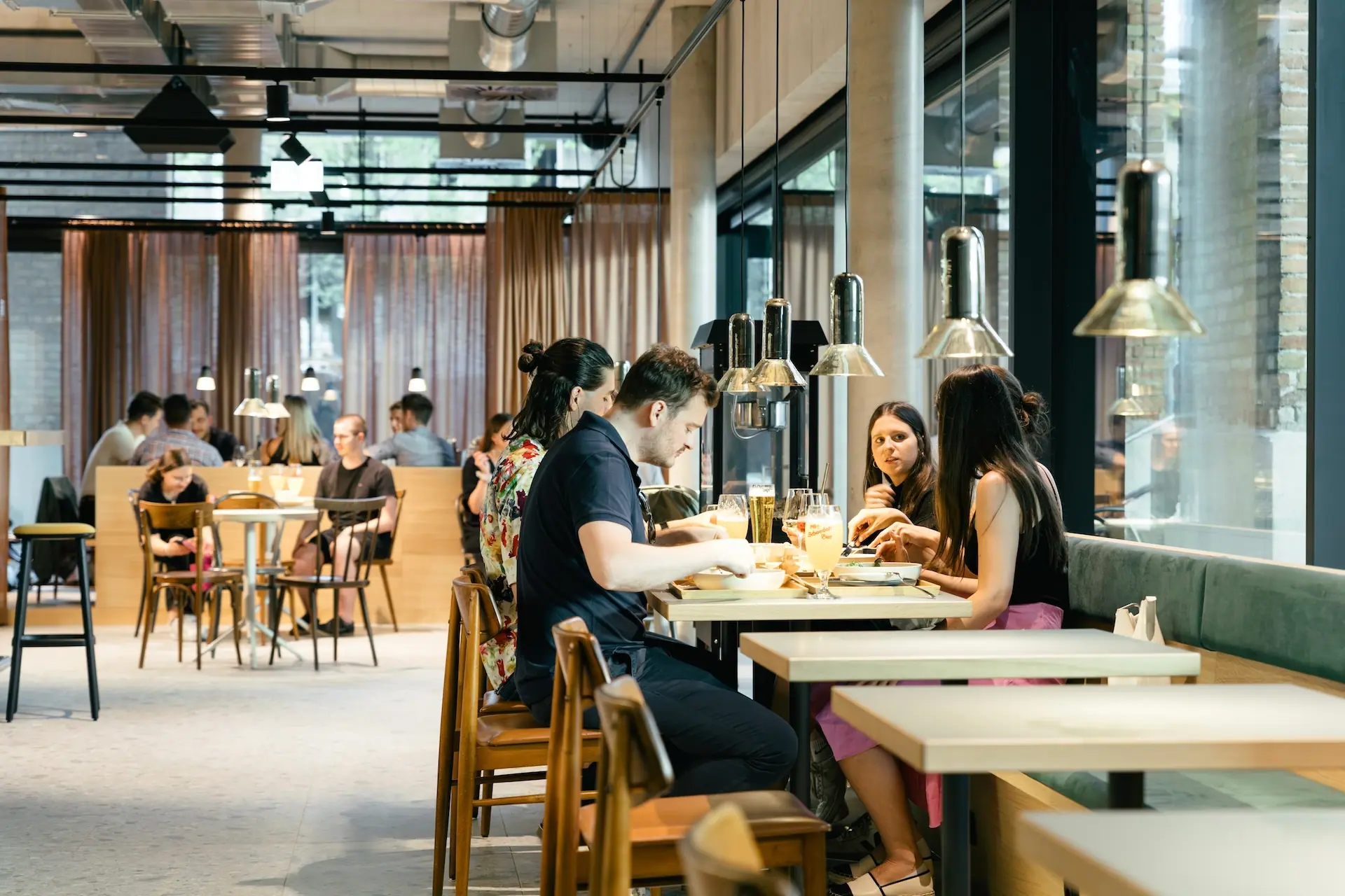 In einem modernen, gut beleuchteten Restaurant mit großen Fenstern und Holzmöbeln wird gegessen und geplaudert. Mehrere Gruppen sitzen an Tischen und genießen das Essen und die Gespräche in einer entspannten Atmosphäre.