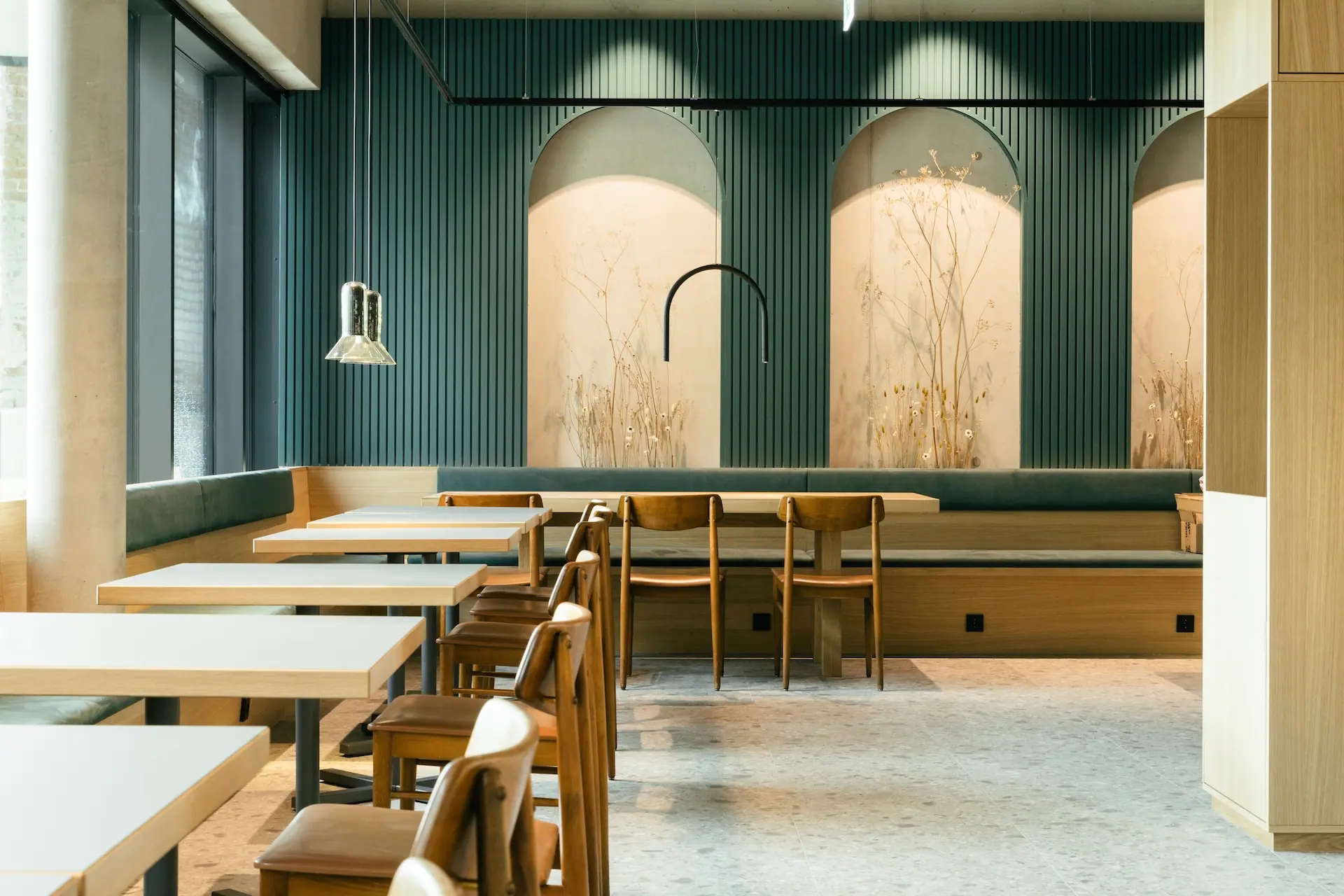 Modernes Café-Interieur mit Holztischen und -stühlen, grün gepolsterten Sitzbänken und dekorativen Wandpaneelen mit botanischen Motiven, die von gewölbten Nischen eingerahmt werden; weiches natürliches Licht strömt durch große Fenster.