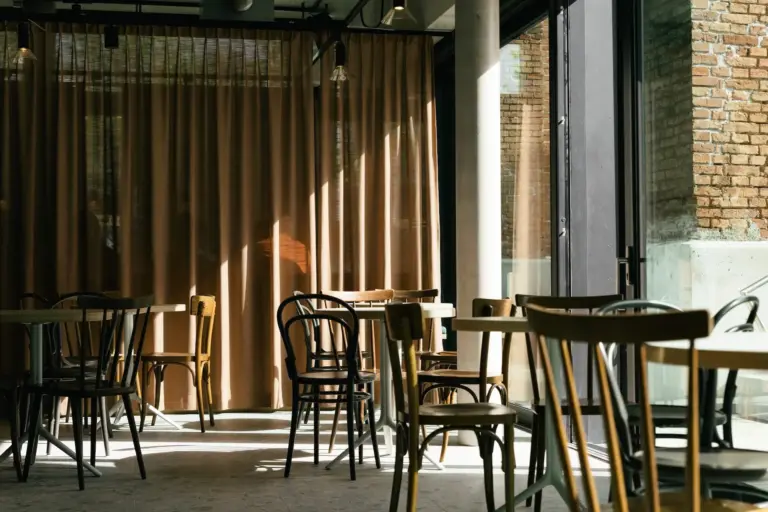 Sonnenlicht strömt in ein ruhiges Café mit leeren Holzstühlen und -tischen. Hellbraune Vorhänge verdecken einige Fenster, und teilweise sind freiliegende Backsteinwände zu sehen. Die Atmosphäre ist ruhig und einladend.
