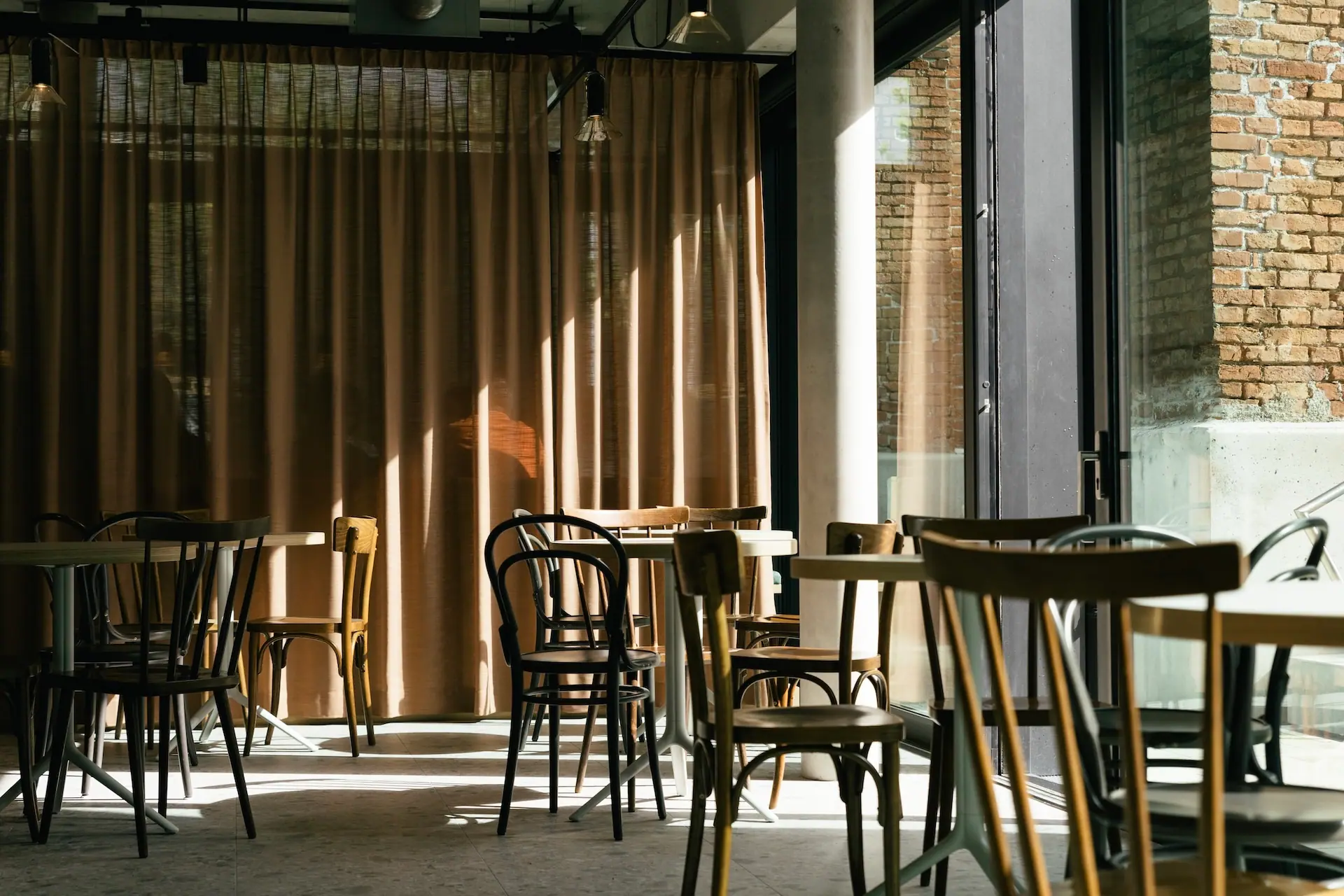 Sonnenlicht strömt in ein ruhiges Café mit leeren Holzstühlen und -tischen. Hellbraune Vorhänge verdecken einige Fenster, und teilweise sind freiliegende Backsteinwände zu sehen. Die Atmosphäre ist ruhig und einladend.