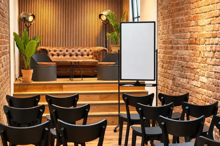 Eine Reihe schwarzer Stühle, die auf eine kleine Bühne ausgerichtet sind, auf der ein braunes Ledersofa, Topfpflanzen und eine leere Whiteboard-Tafel stehen.