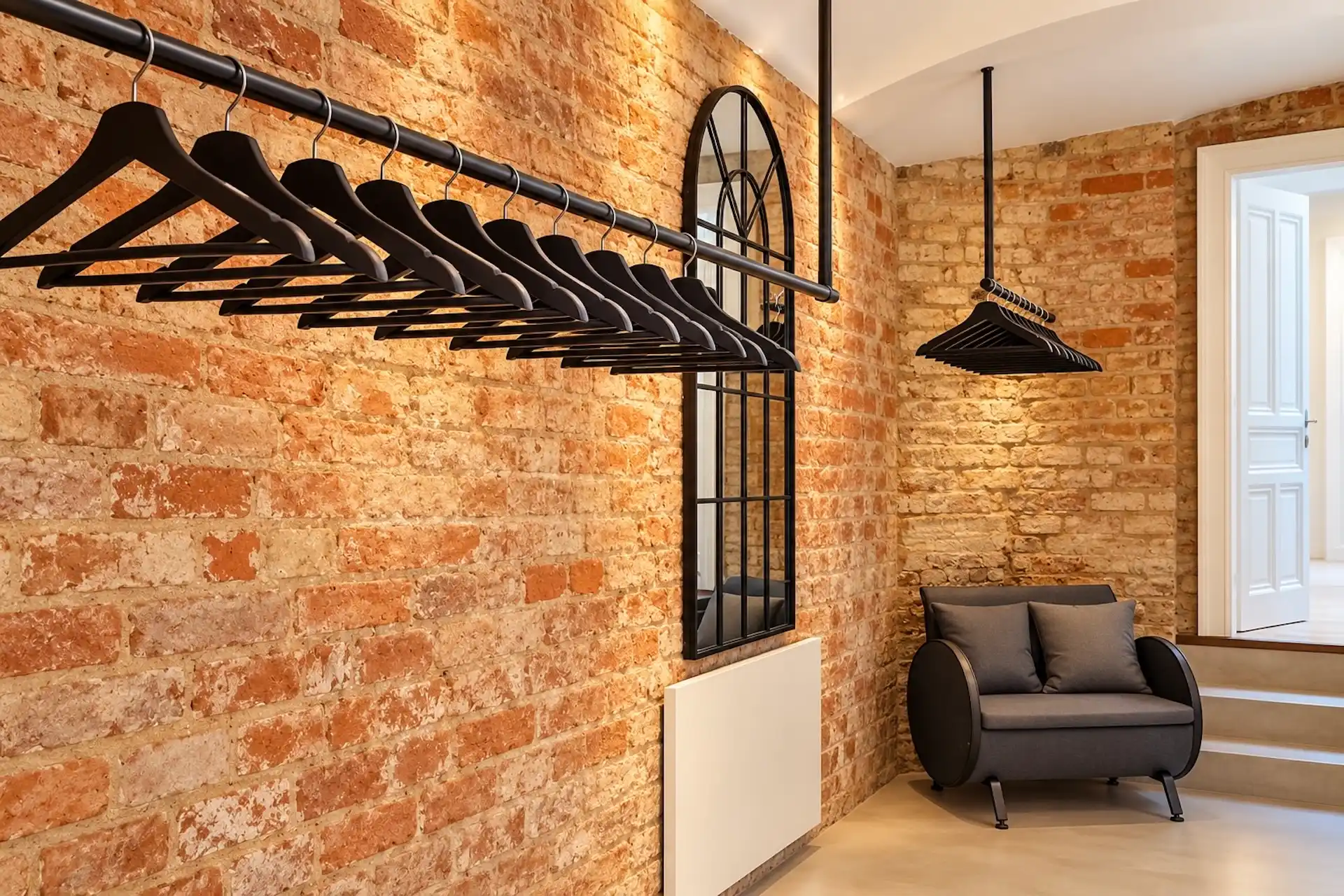 Leere schwarze Kleiderbügel hängen an zwei Schienen im Industrial-Stil vor einer unverputzten Backsteinwand; in der Ecke stehen ein gewölbter Spiegel und ein grauer Sessel.