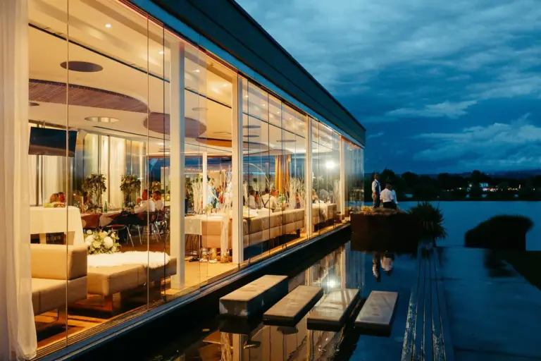 Ein modernes gläsernes Gebäude leuchtet in der Abenddämmerung warm auf und gibt den Blick frei auf eine lebendige Veranstaltung im Inneren. Draußen kreuzen Trittsteine einen spiegelnden Pool mit einem dramatischen, wolkenverhangenen Himmel im Hintergrund.